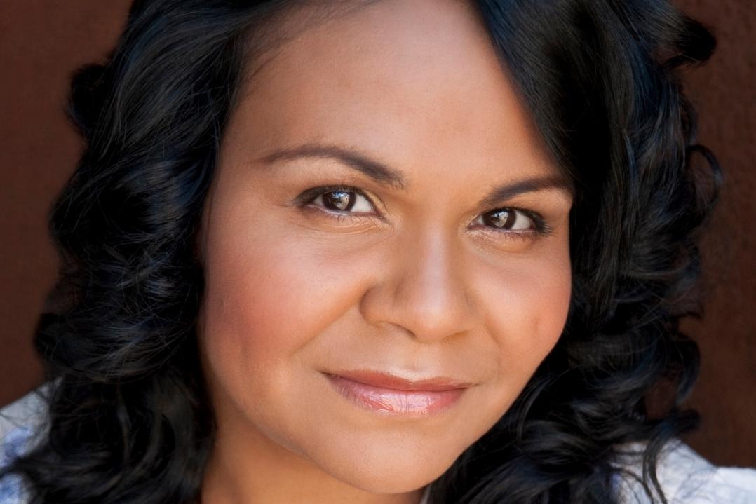 Smiling woman with dark, wavy hair against a dark background. Her expression is calm and friendly, conveying warmth and approachability.