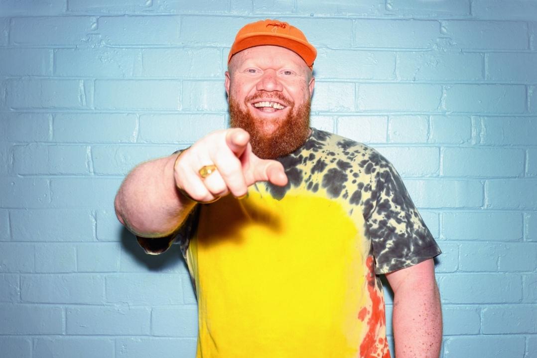 Smiling man in a bright outfit points playfully at the camera. He wears a yellow and patterned shirt with an orange cap, standing against a blue brick wall.