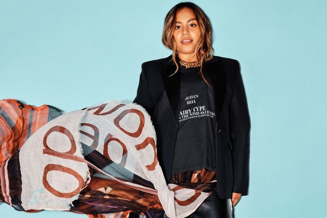 A person in a black blazer and t-shirt smiles against a blue background. A scarf with large ddraped over one shoulder, adding a touch of whimsy.