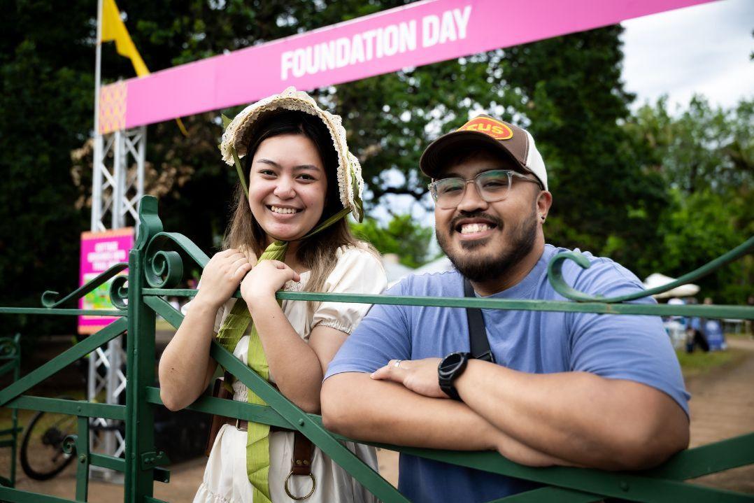 A smiling woman in a floral dress and hat stands beside a man in a blue shirt and cap. They lean on a fence under a pink banner, outdoors, joyful vibe.
