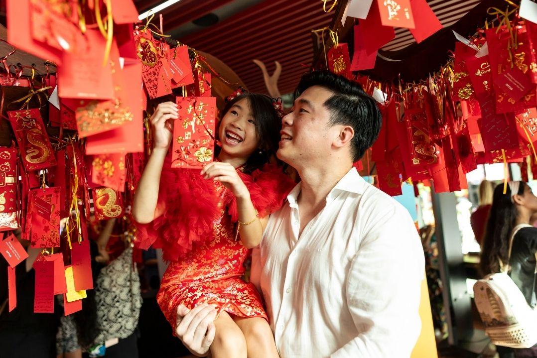 A joyful couple in red attire admires vibrant red lanterns hanging overhead, surrounded by warm light, capturing a festive and celebratory atmosphere.
