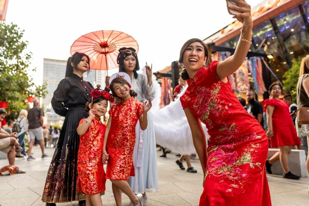 A group of people in vibrant, traditional attire take a joyful selfie outdoors. The scene conveys celebration and cultural pride on a busy street.