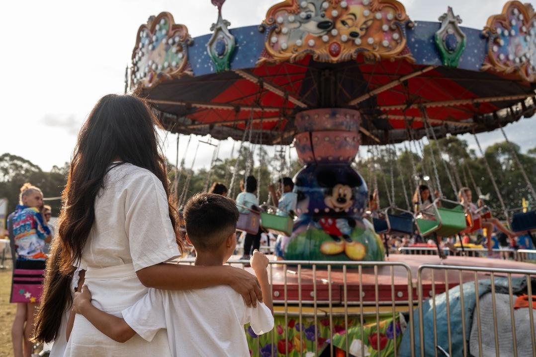 A woman and child, seen from behind, stand in front of a colourful carousel at an amusement park. They appear relaxed, enjoying a lively, cheerful atmosphere.