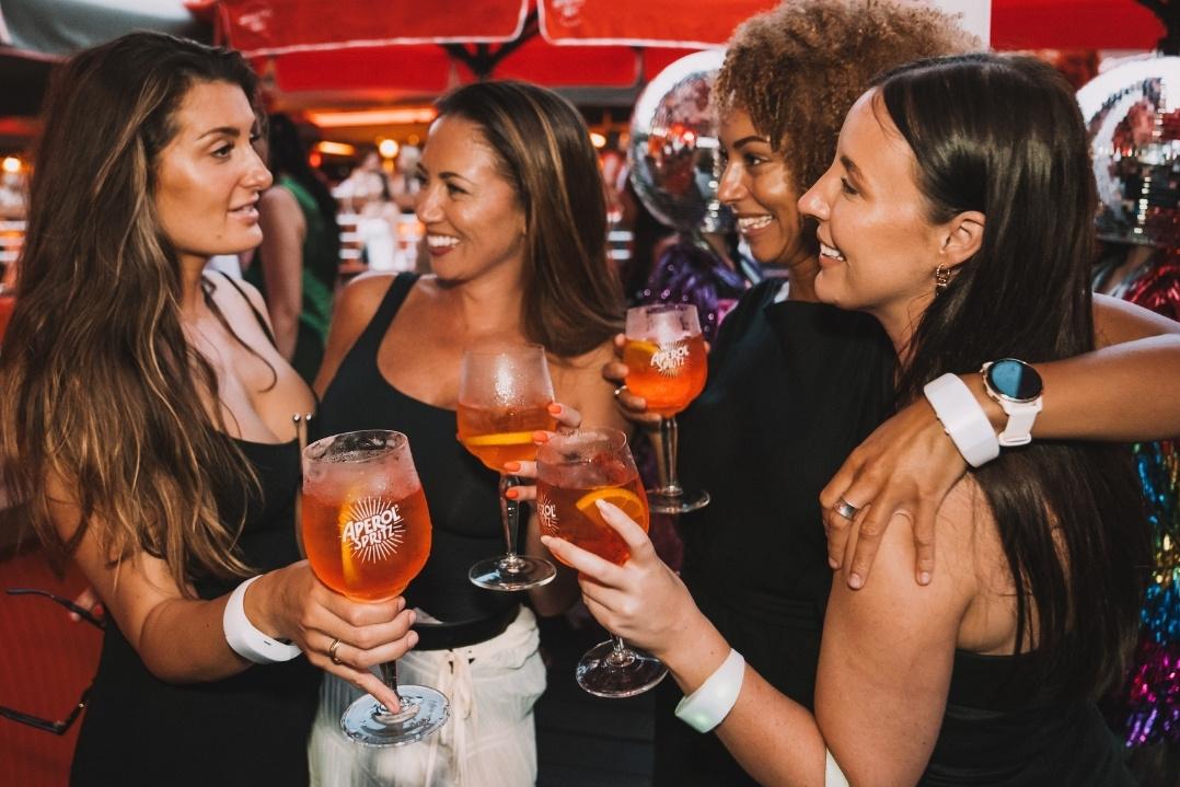 Four women in a lively bar scene clink glasses filled with a bright orange drink, smiling and laughing, conveying a festive and joyful atmosphere.