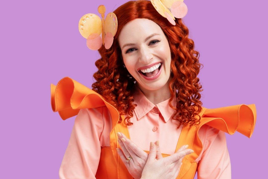 Woman with curly red hair wearing a vibrant orange and pink costume adorned with flowers, smiling joyfully against a white background. Whimsical and cheerful.