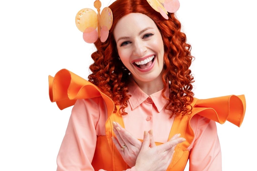 Woman with curly red hair wearing a vibrant orange and pink costume adorned with flowers, smiling joyfully against a white background. Whimsical and cheerful.