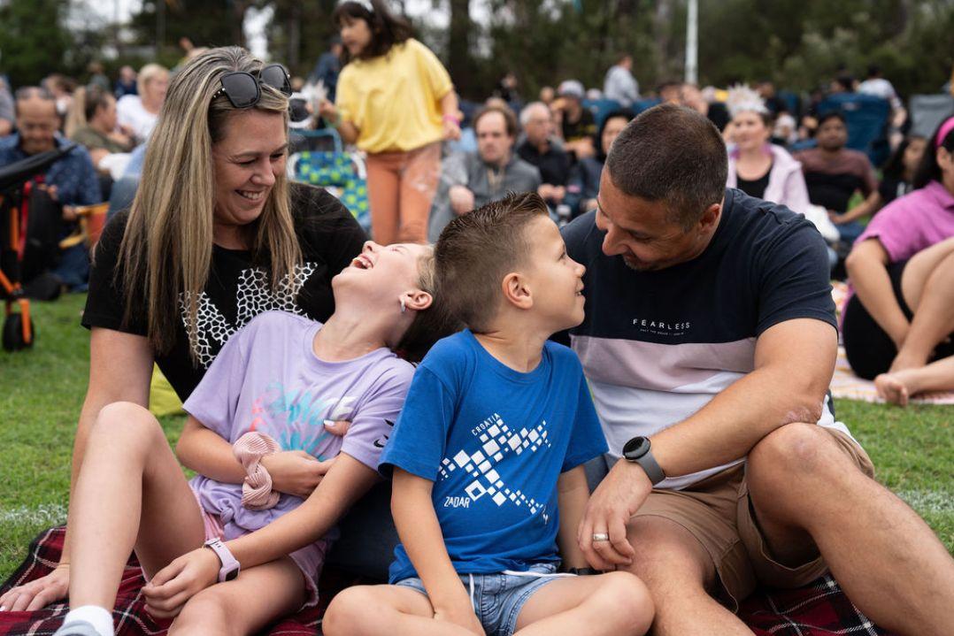 A family of four sits on a blanket in a park, smiling and gazing at each other. The setting is a lively outdoor gathering with people in the background.