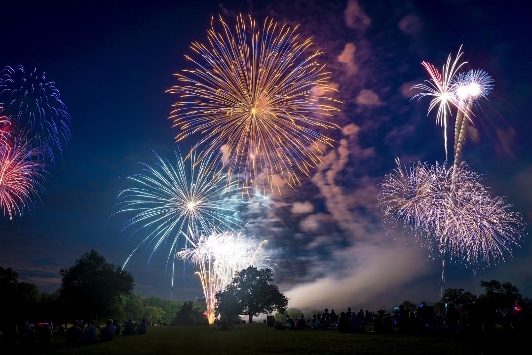 A vibrant fireworks display lights up the night sky with bursts of red, orange, and blue over silhouetted trees, creating a festive and celebratory atmosphere.