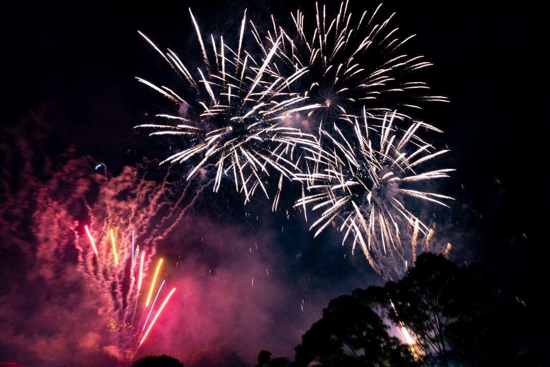Bright fireworks light up a night sky, with bursts of white and pink hues. Silhouetted crowd watches below, creating a festive and awe-inspiring scene.