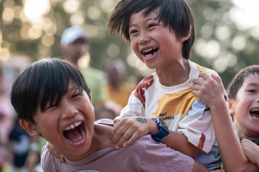 Two children in a joyful moment outdoors. The one in front gives a piggyback ride to the other, both laughing. The background features blurry trees and people.