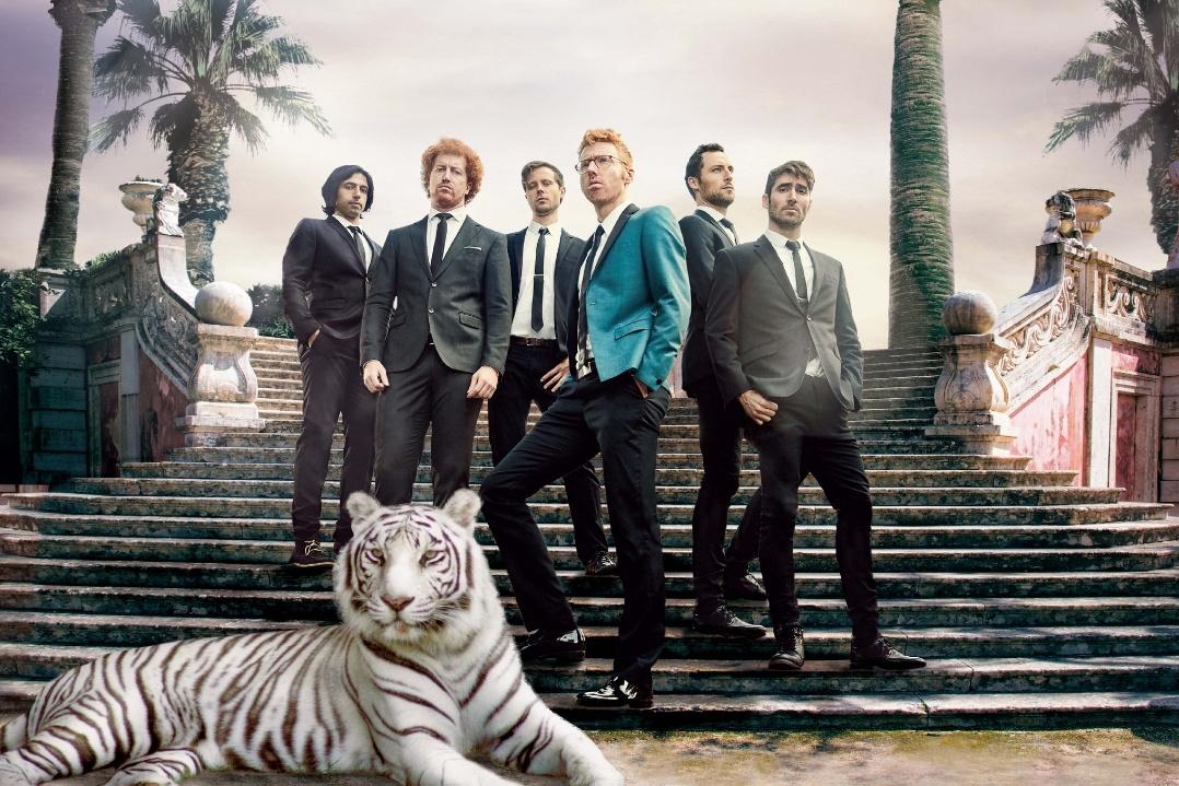 A group of men in formal suits stands on grand stone steps with palm trees in the background. A calm white tiger lies in the foreground. The mood is elegant and stylish.
