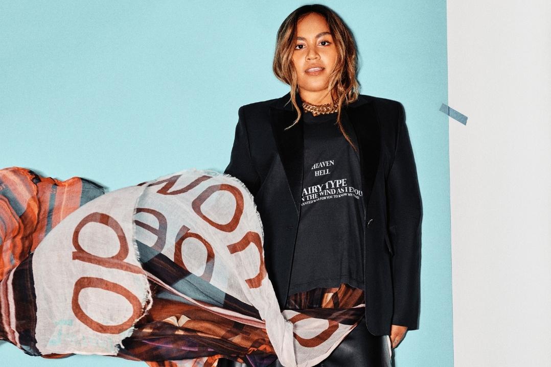 A person in a black blazer and shirt smiles warmly against a light blue background.