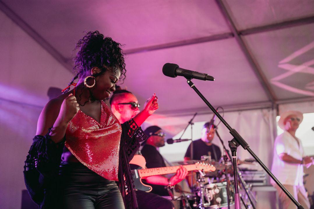 A lively band performs on a stage under purple lighting. The lead singer, in a red outfit, sings passionately. Drummer and guitarist play energetically.