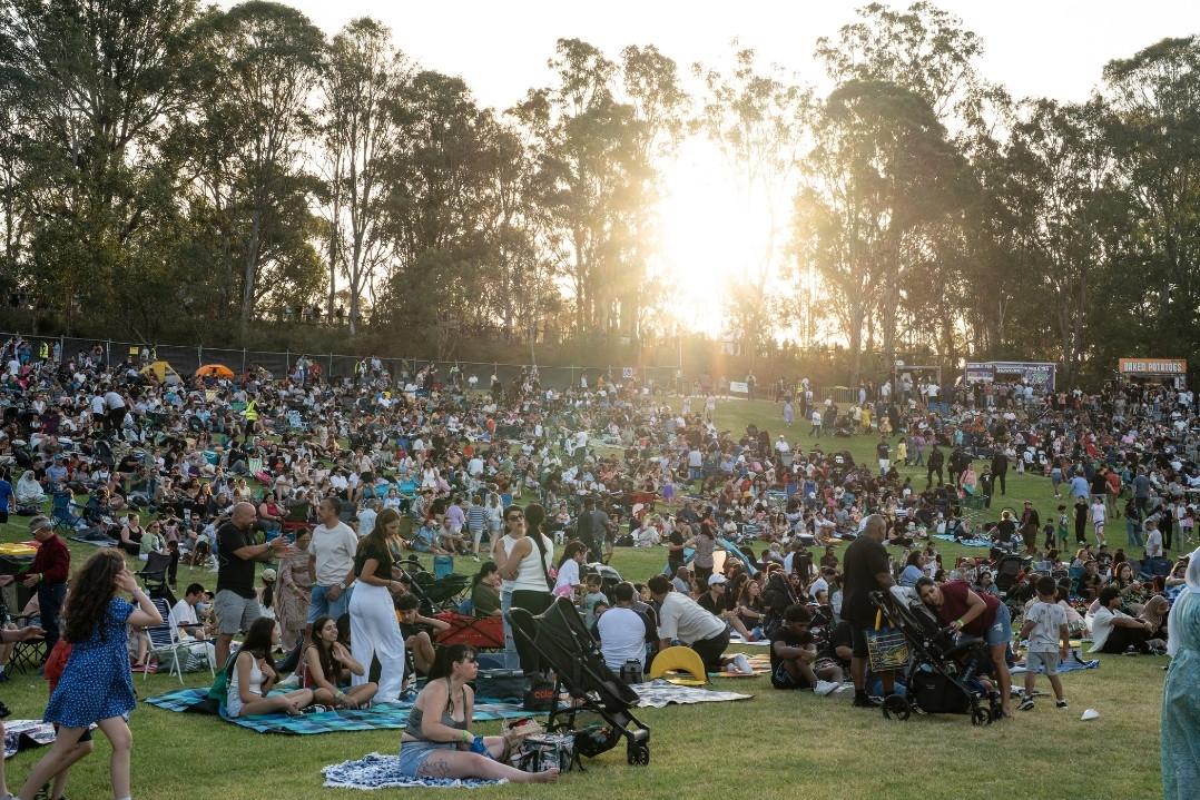 A large crowd gathers outdoors on a sunny day, sitting on grass and blankets, with sunlight filtering through trees, creating a warm, festive atmosphere.