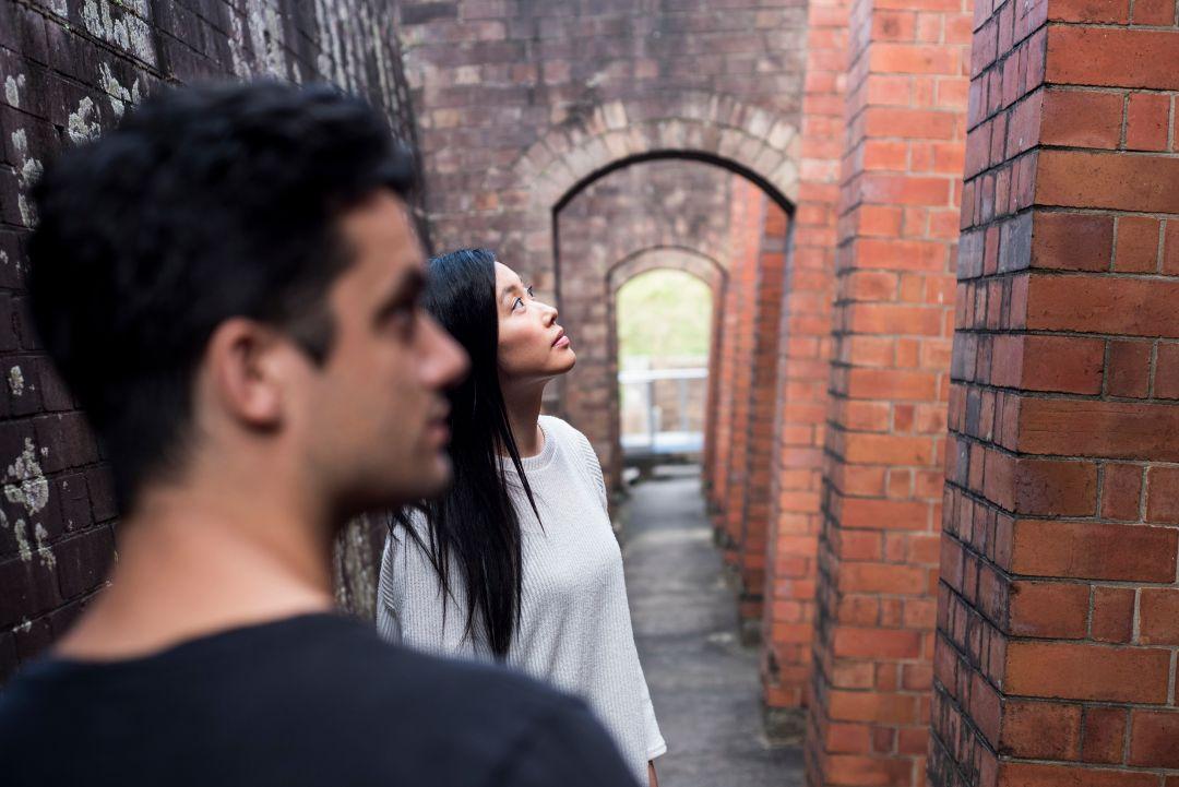 A man and woman stand in a narrow brick alley, gazing upward. The scene conveys curiosity and interest, with warm sunlight filtering through.