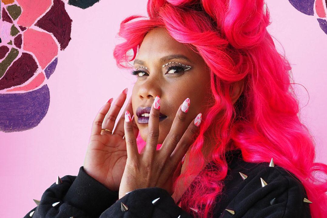 A person with vibrant pink hair poses against a floral pink background. They wear a black jacket adorned with metallic accents, with a surprised expression.