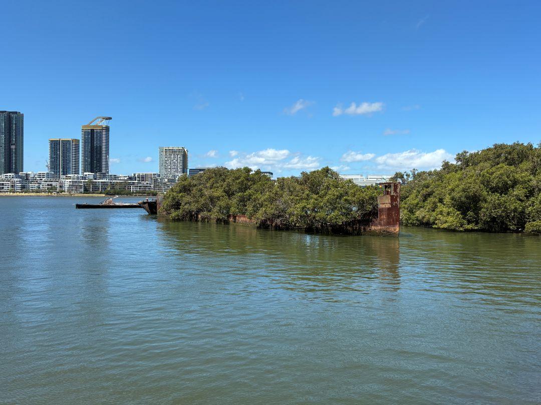 Calm river scene with lush green islands in the foreground, set against a backdrop of modern skyscrapers under a clear blue sky, evoking tranquility.