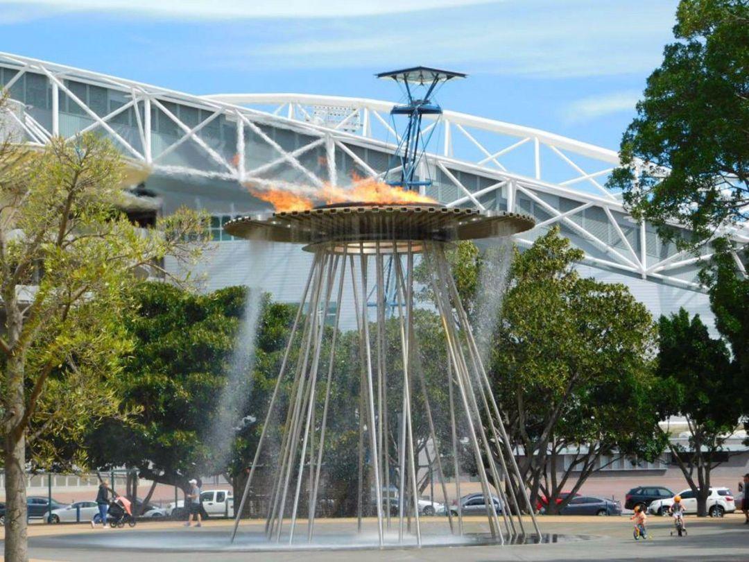 A modern fountain with a circular canopy and tall, thin metal supports, framed by leafy trees and a white bridge under a blue sky, conveys a serene urban scene.