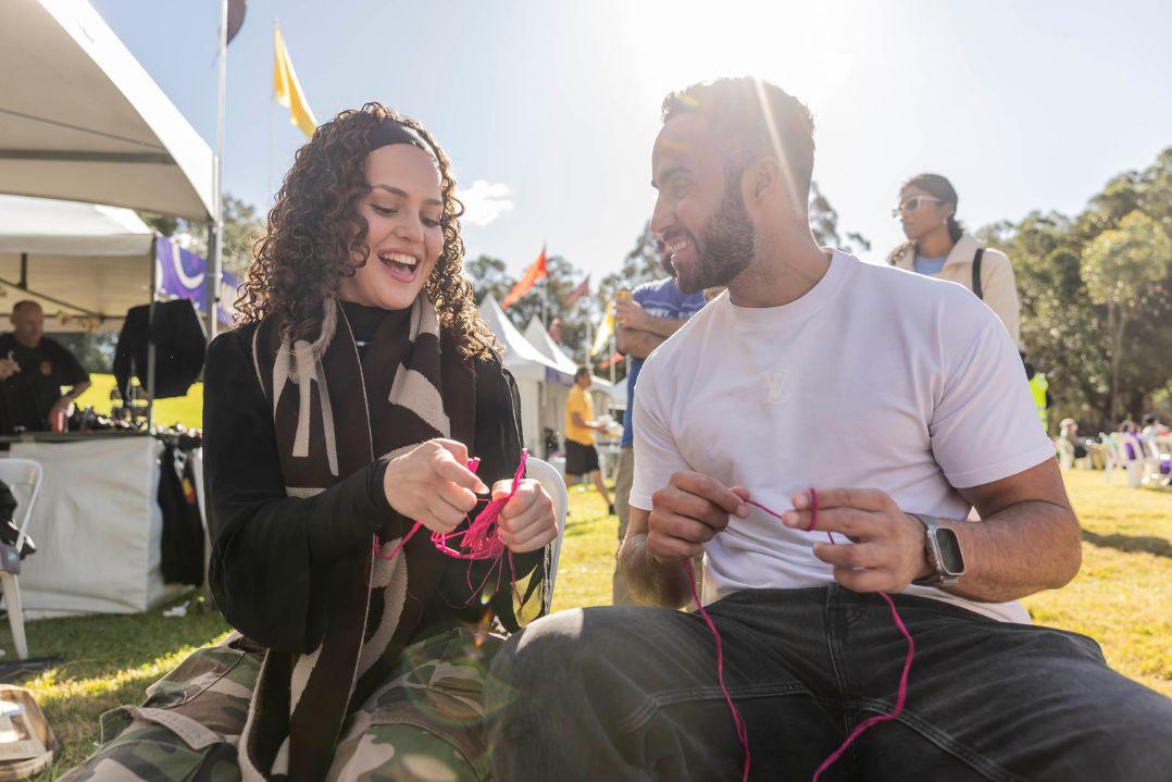 A woman in a black outfit and a man in a white shirt are outdoors, smiling while painting on a sunny day. People and a tent are visible in the background.