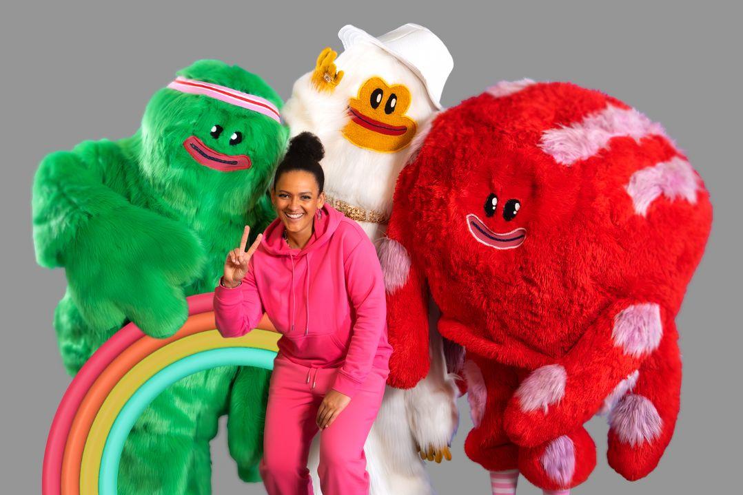 A person in a pink tracksuit smiles brightly, surrounded by large, colourful furry creatures—green, red, and white with cheerful expressions. Playful and whimsical vibe.