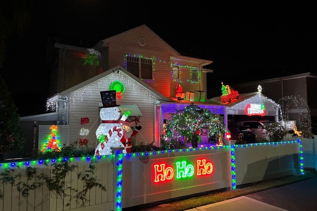 Festive house decorated with bright Christmas lights, featuring a glowing "Ho Ho Ho" sign, Santa, snowman, and reindeer inflatables on the lawn.