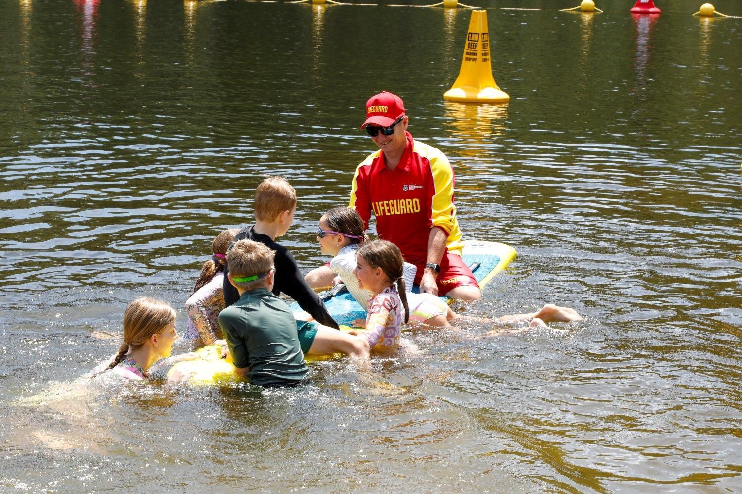 A lifeguard in red oversees two children playing with a green kayak in a lake. The scene is lively and joyful, with bunting in the background.