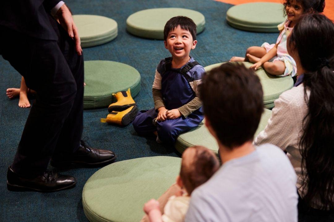 A young child sits laughing on the floor among green cushions, surrounded by adults and children. The scene is joyful and interactive.