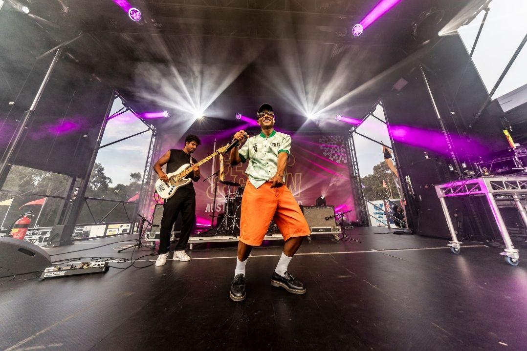 A dynamic stage performance with a male singer in orange shorts, animatedly holding a mic. A guitarist plays behind, with vibrant purple stage lights.