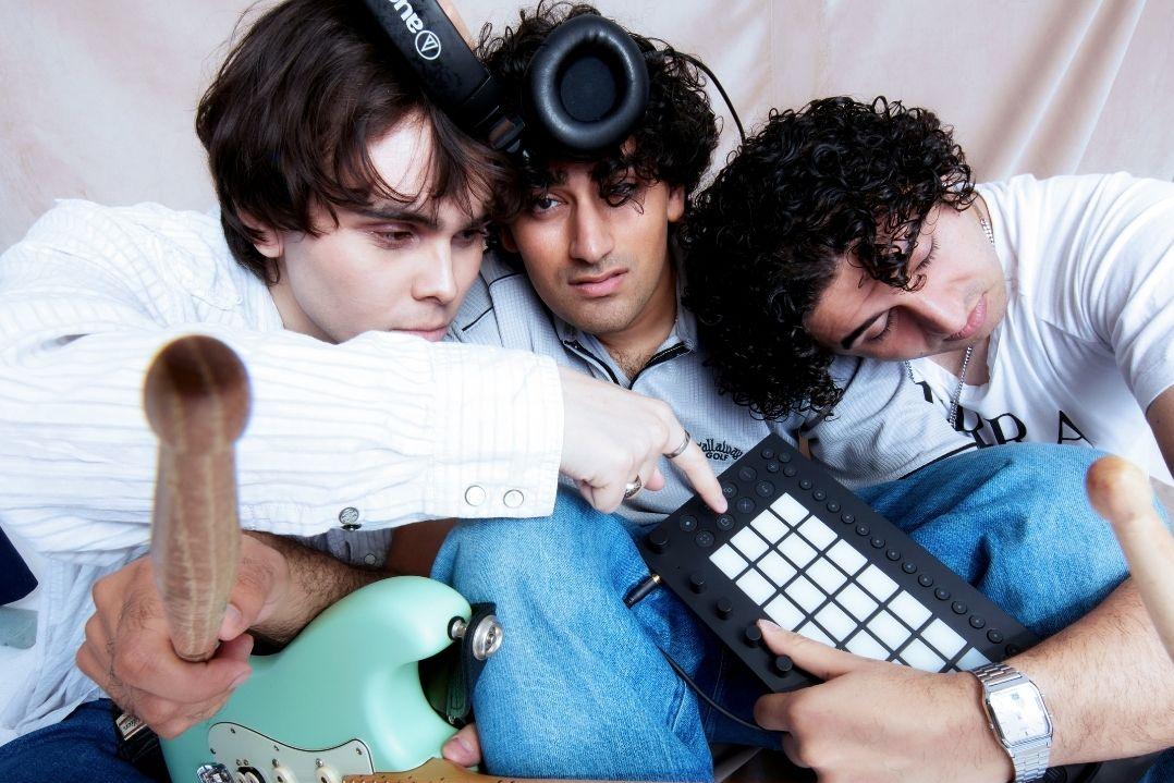 Three people in casual clothing sit closely together, surrounded by musical instruments including a guitar and drum pad, conveying a playful and creative mood.