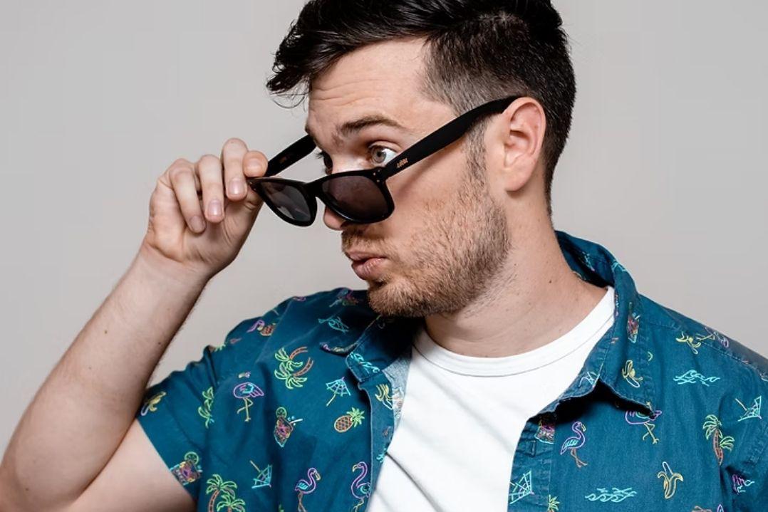A man in a blue floral shirt lifts his sunglasses with a curious expression. The neutral background highlights his casual, inquisitive demeanour.