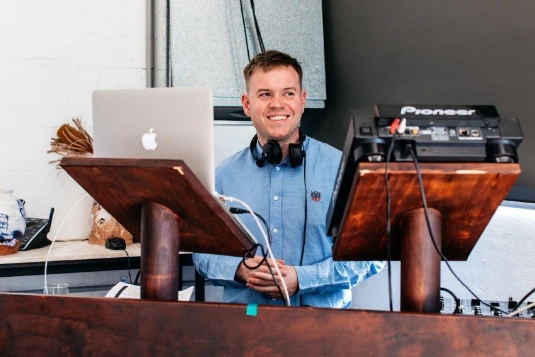 Smiling man in blue shirt stands at a wooden music stand with a laptop and audio equipment, wearing headphones, conveying a joyful and focused vibe.