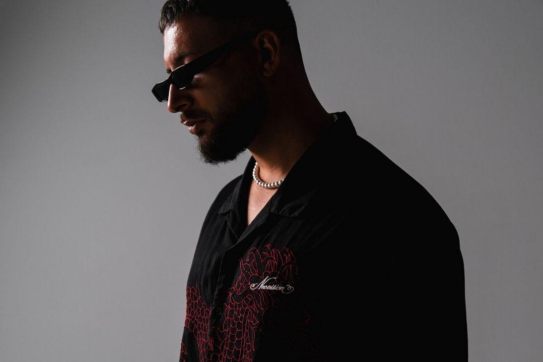 A man in sunglasses and a dark shirt stands in shadow against a gray background, creating a mysterious and contemplative mood.