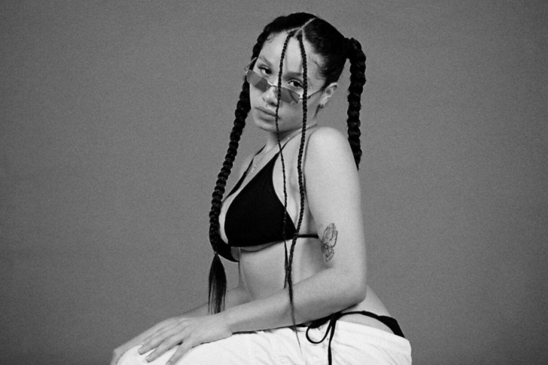 A woman with long braided hair poses in a black bikini against a grey background. Her expression is serene and thoughtful, creating a calm and elegant mood.