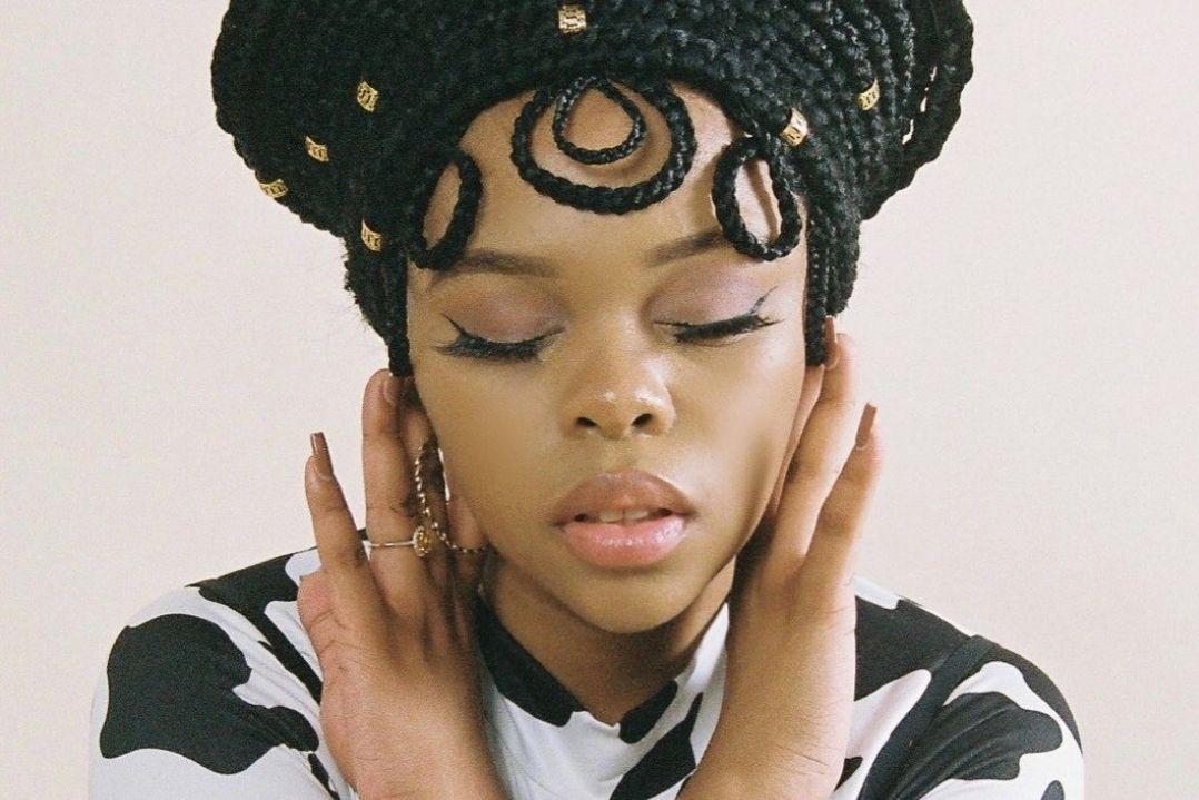 Woman with intricate braided hairstyle and patterned top, eyes closed, gently touching her neck. The expression is serene and contemplative.