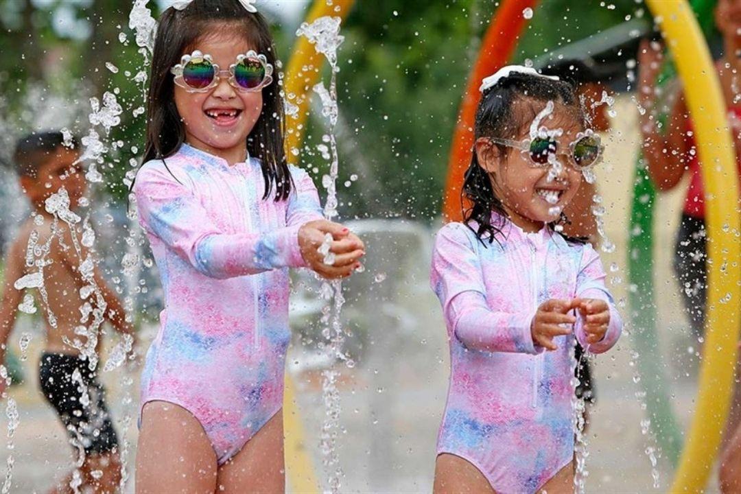 Two young children in swimsuits joyfully play with water jets at a splash park, wearing goggles and smiling under colourful arches. Sunny day, vibrant scene.