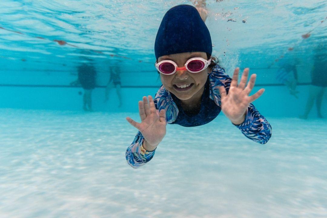 A child in red goggles and a navy cap swims underwater in a clear pool. Their expression appears playful and focused amidst shimmering blue water.
