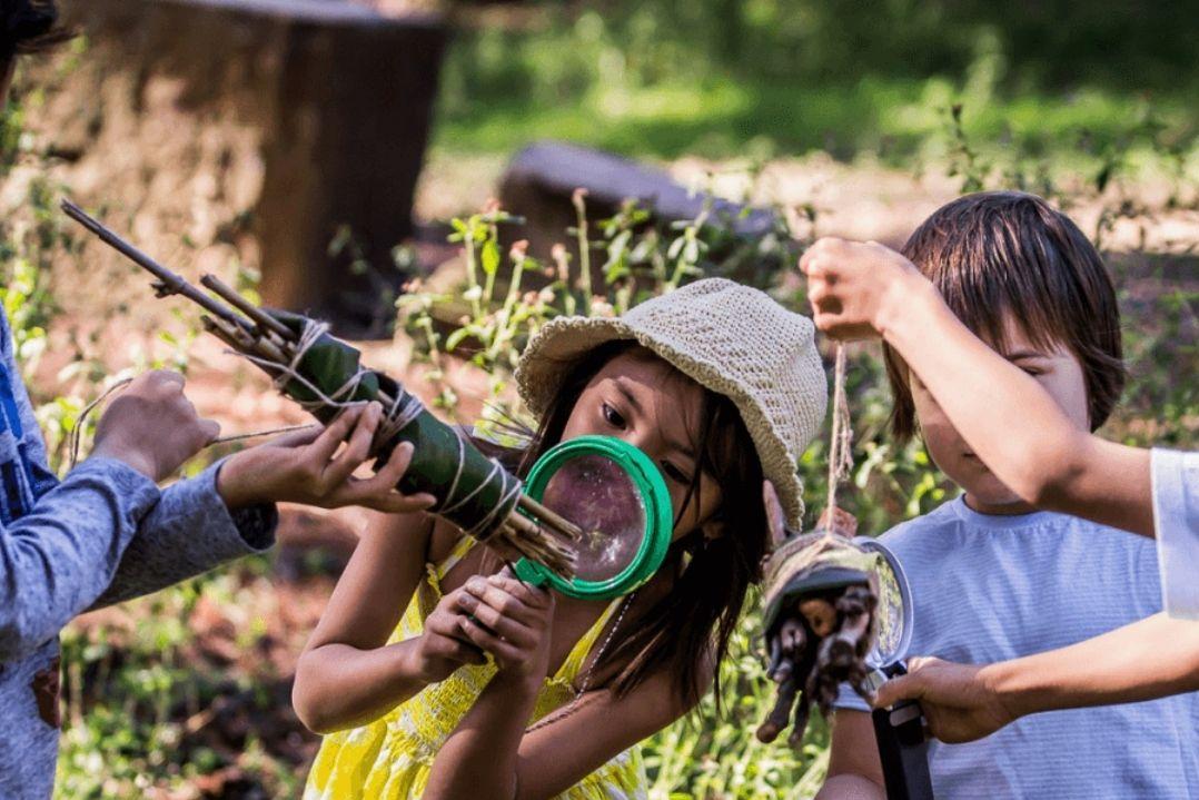 Children joyfully play outdoors, each using natural materials to make creative horn shapes. The setting is lush and green, capturing a sense of fun and imagination.