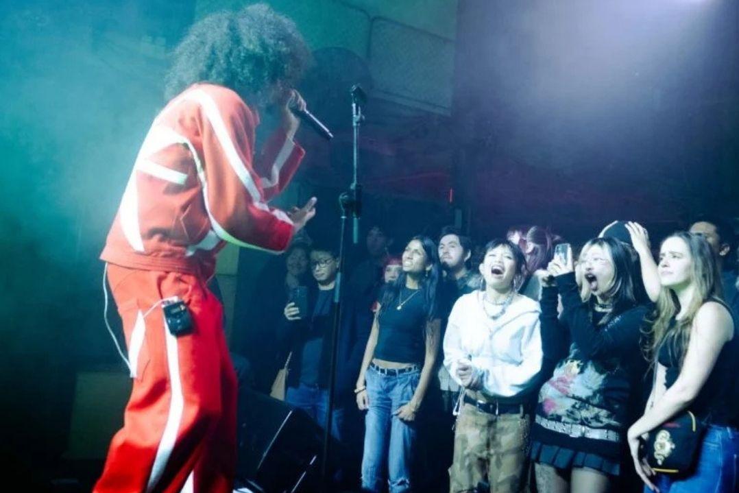 Performer in a red and white outfit energetically sings on stage, engaging an enthusiastic crowd under vibrant blue and purple stage lights.