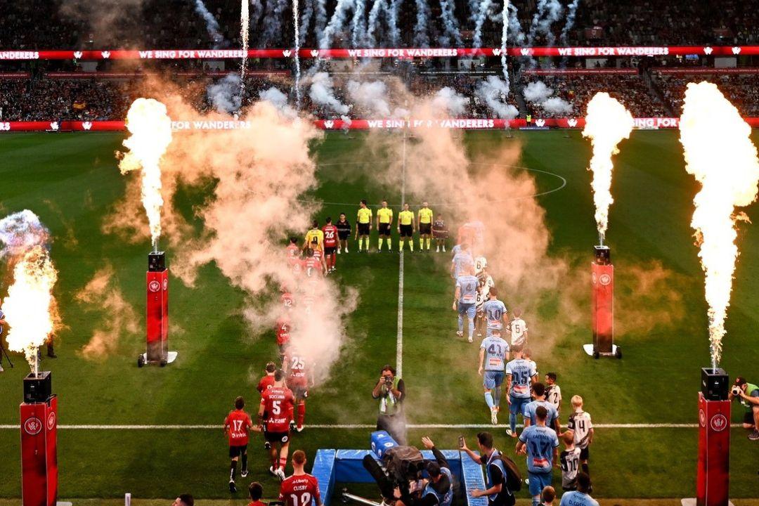 Teams enter a stadium through fireworks and smoke, creating an exciting atmosphere. The scene is vibrant, celebrating the start of a sports event.