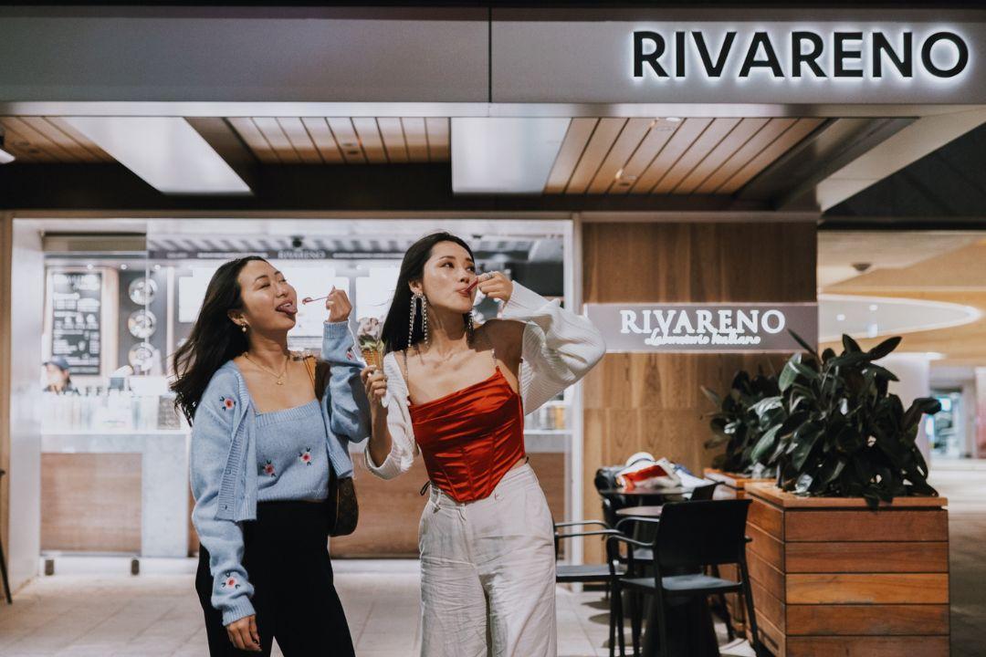 Two women happily walk inside a modern Rivareno gelato shop. One holds a cone, wearing white pants and a red top; the other wears black pants and a blue top.