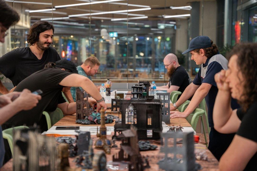 A group of people sit around a table covered with various bottles and glasses, engaged in lively conversation and tasting. The atmosphere is casual and social.