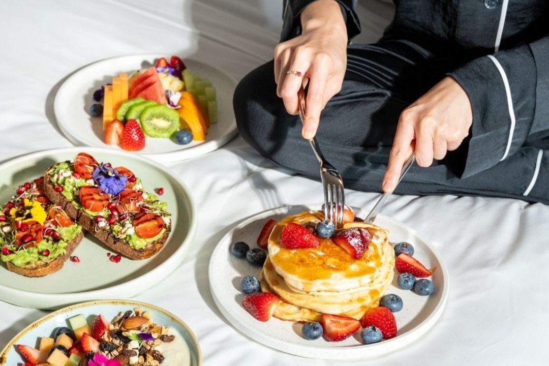 A person in dark clothing prepares to eat pancakes topped with berries and flowers on a bed. Nearby are colourful plates of waffles and avocado toast.