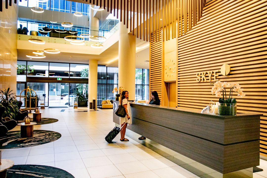 Modern hotel lobby with warm lighting and wooden accents. A person checks in at a sleek reception desk while another lounges on a sofa, conveying comfort.