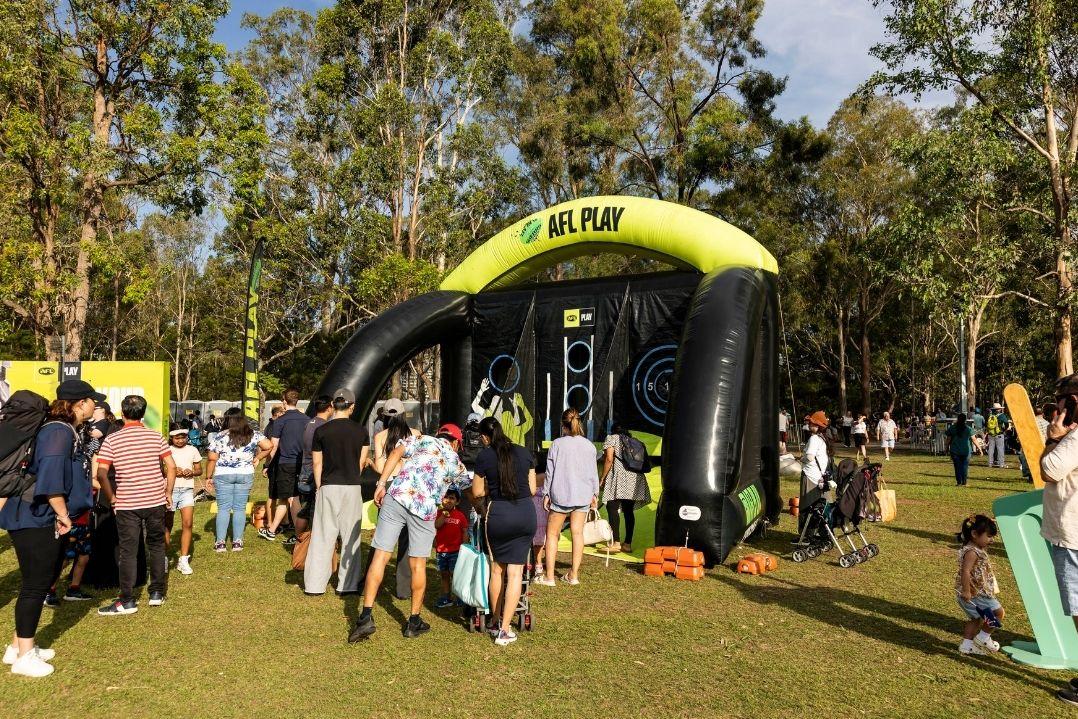 People gather in a sunny park near an inflatable archway that reads "AFL." Trees in the background and a festive, cheerful atmosphere.