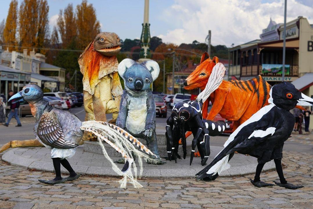 A lively outdoor scene depicts people wearing large, colourful animal masks and costumes. The atmosphere is festive, with a backdrop of autumn trees and buildings.