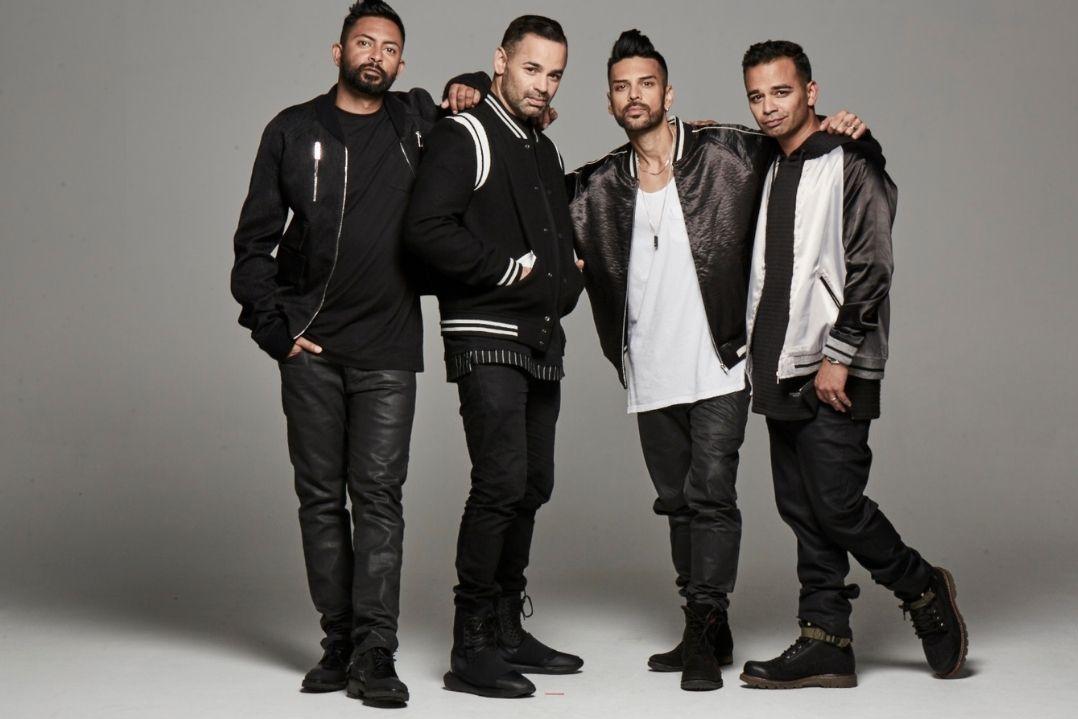 Four men stand closely together against a gray background, wearing casual, modern outfits. They project a sense of camaraderie and confidence.