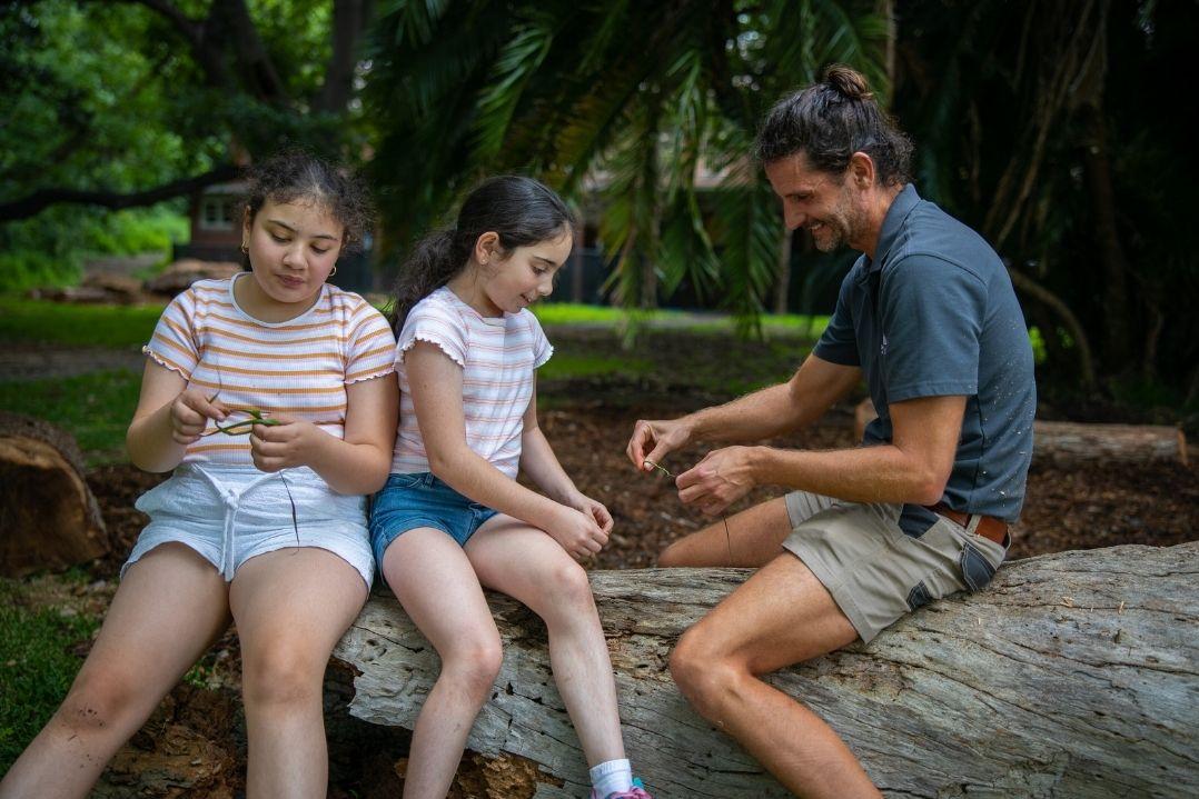 A man and two young girls sit on a log in a forest, crafting with twine. The girls look focused, the setting is lush and green, creating a peaceful, engaging mood.