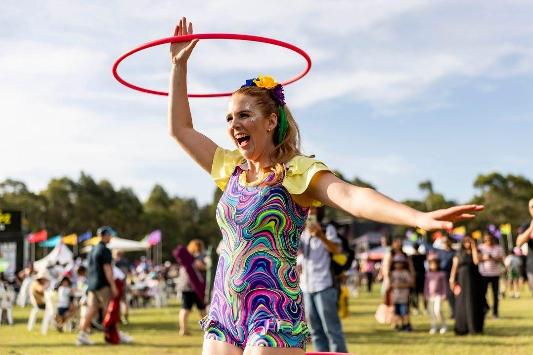 A woman joyfully spins a red hula hoop above her head at an outdoor festival. She wears a colourful dress, surrounded by a lively crowd under a sunny sky.