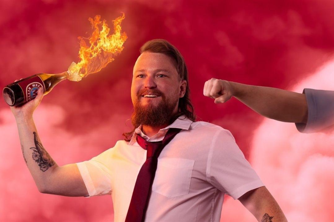 A person with medium-length hair and a beard holds a flaming Molotov cocktail, wearing a shirt and tie against a red smoke-filled backdrop, looking intense.