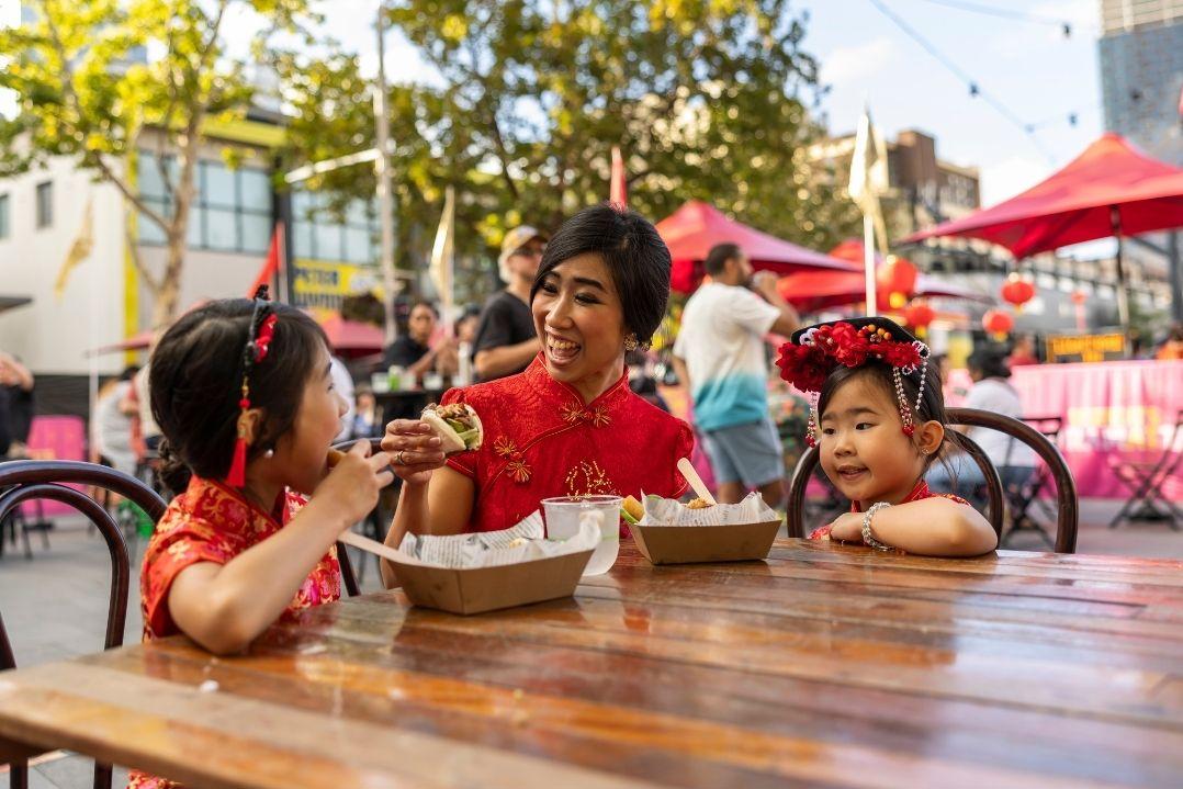 A woman and two kids enjoy ice cream at an outdoor café. They smile and chat at a wooden table under trees and red umbrellas, creating a joyful atmosphere.